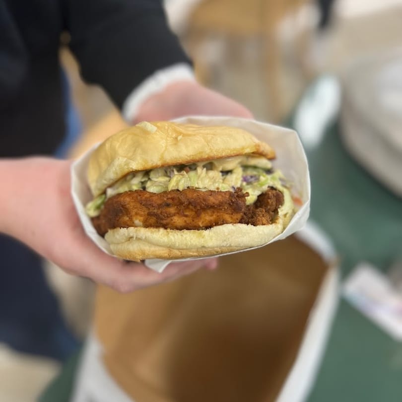Hands holding a Green Chile House fried chicken sandwich with coleslaw in a paper wrapper.
